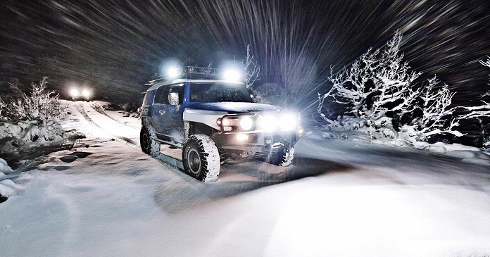 4x4 white and blue SUV in snow with turned on pedestal-mounted and flush-mounted led lights on a mountain at night.