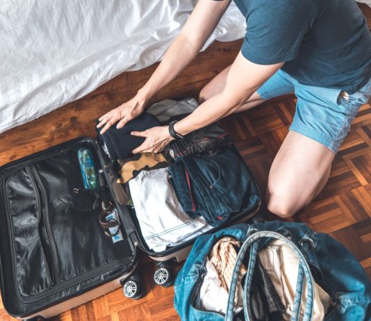 Person packing clothes into a suitcase next to a bed, preparing for travel.