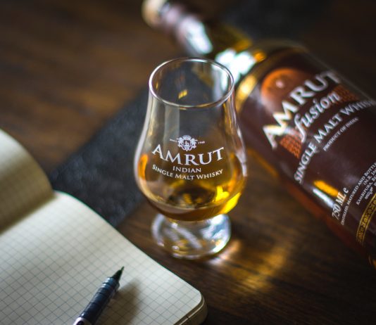 Amrut Indian single malt whisky with a glass and a notebook on a wooden table.